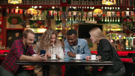 Afro-American-man-with-friends-in-a-cafe-laughs-and-spends-time-in-a-cheerful-company-with-a-girl-watching-photos-and-smiling-while-telling-stories.